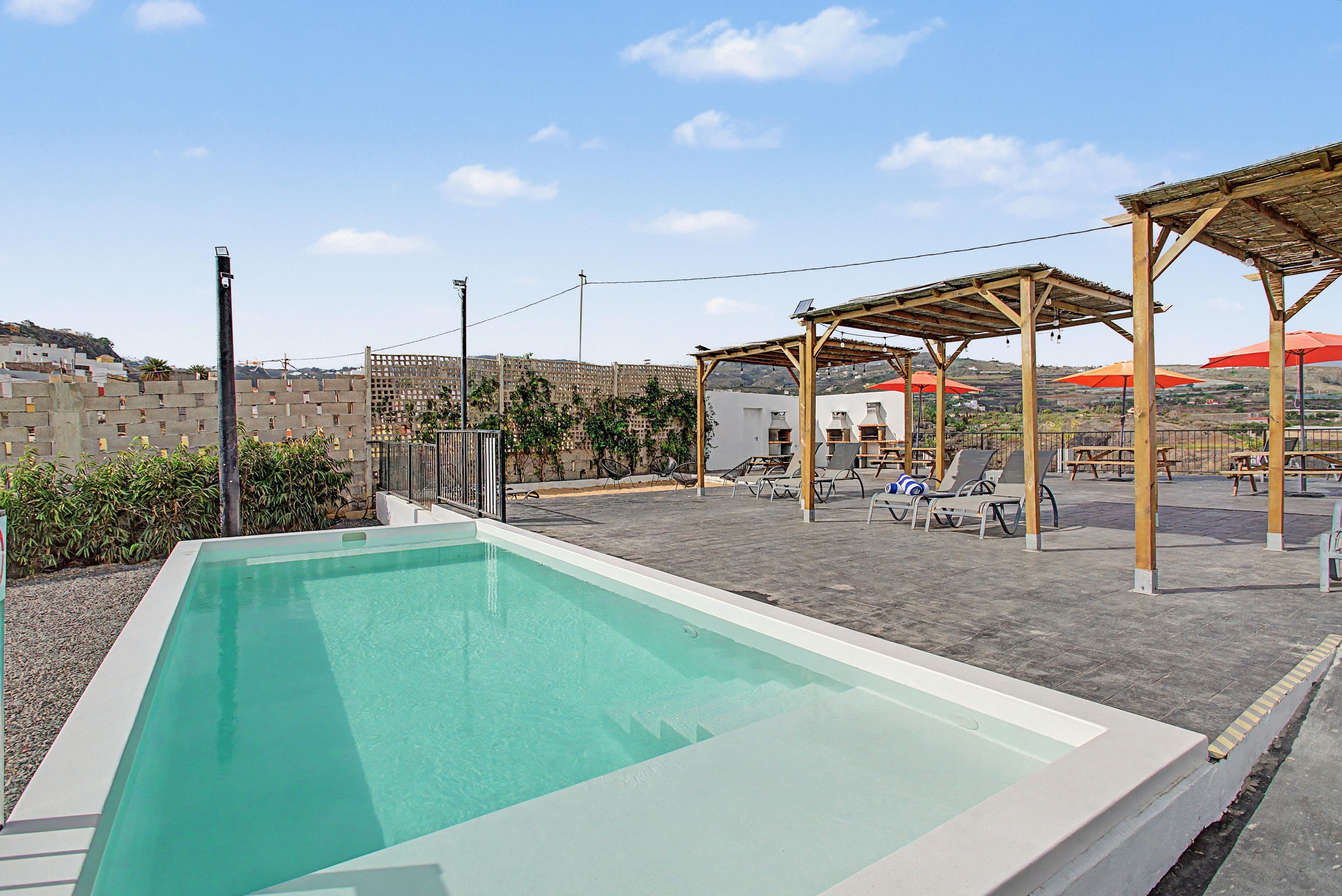 A rectangular swimming pool with a clear blue water surface, surrounded by a paved patio area with lounge chairs and a pergola structure.