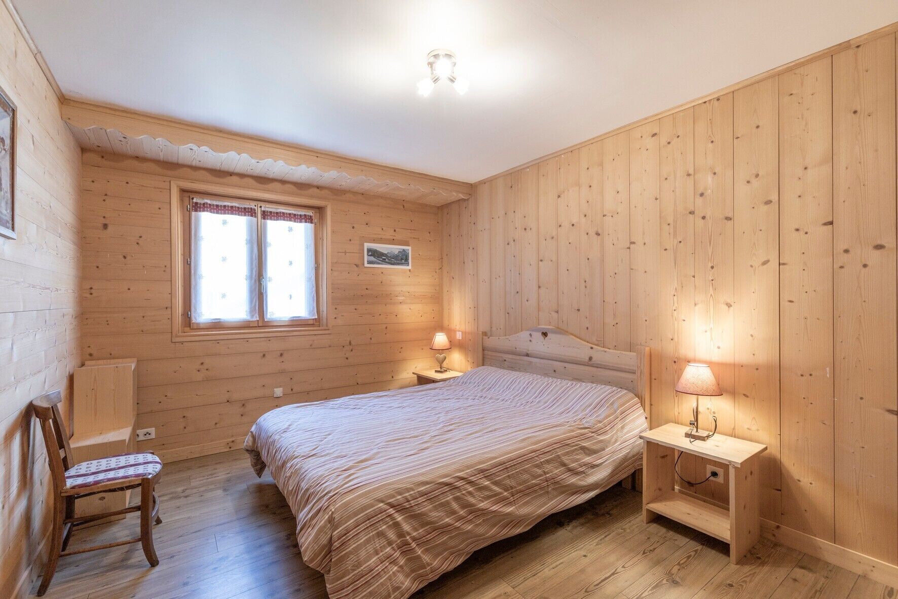 A wooden-paneled bedroom with a bed, two bedside tables, a chair, and a window with curtains.
