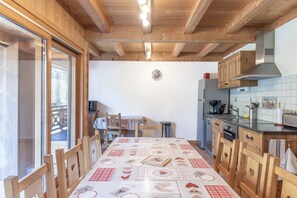Une cuisine avec des meubles de rangement en bois, une table à manger recouverte d’une nappe à carreaux et une horloge au mur.