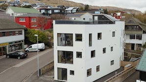 A modern multi-story building with large windows and a flat roof, located in a residential area with houses in the background. - A Varoa Apartments (Torshavn)