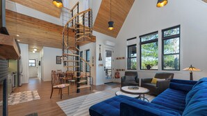Un amplio espacio habitable con un sofá azul, una escalera de madera y grandes ventanales que ofrecen luz natural.