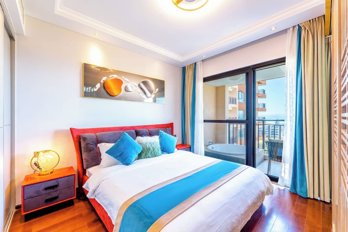 A modern hotel room with a large bed, a nightstand, a wall-mounted artwork, and a balcony with a view of the city.