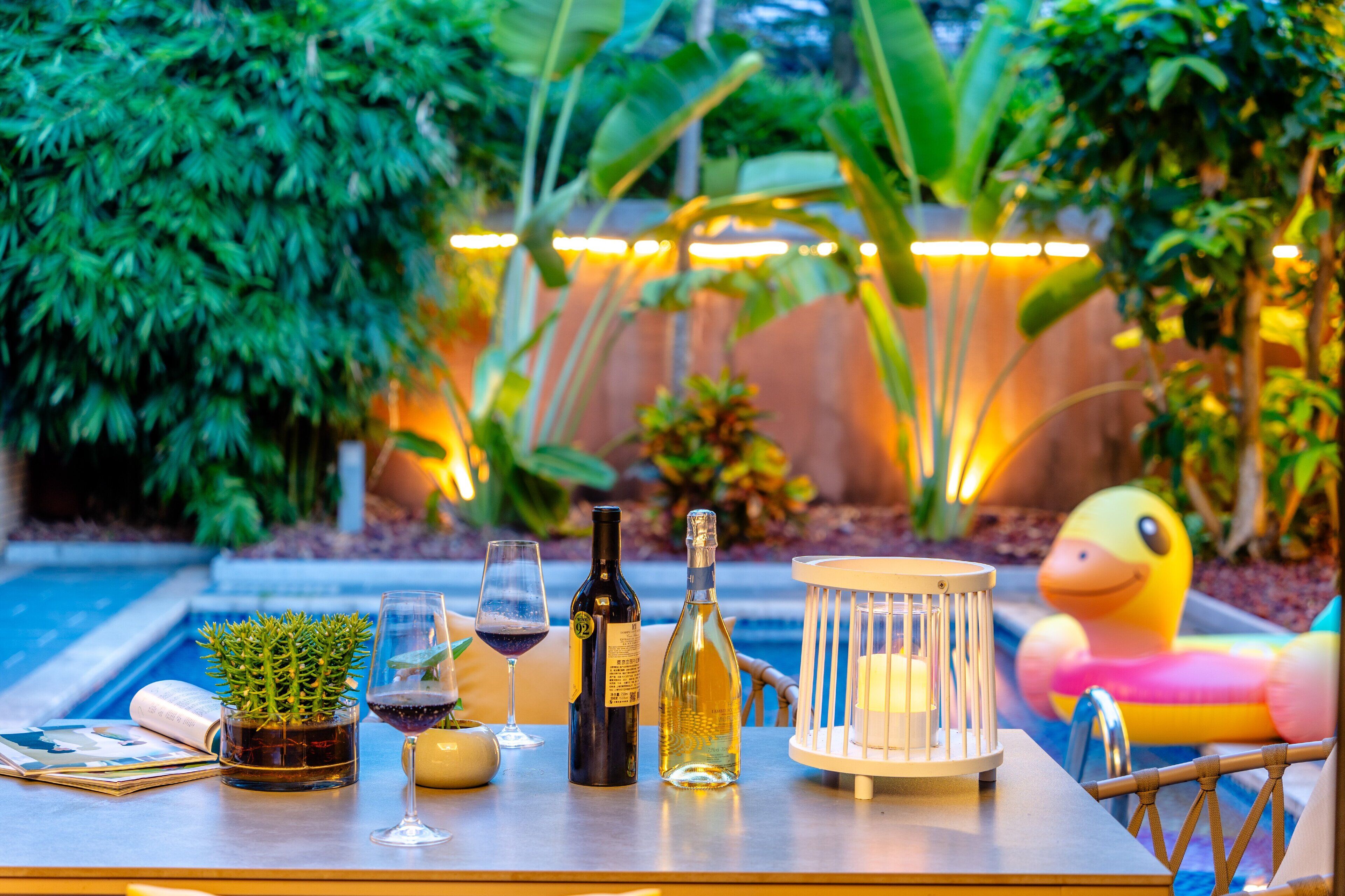 Una mesa junto a la piscina con una botella de vino, una copa de vino, una vela y un flotador con forma de pato.