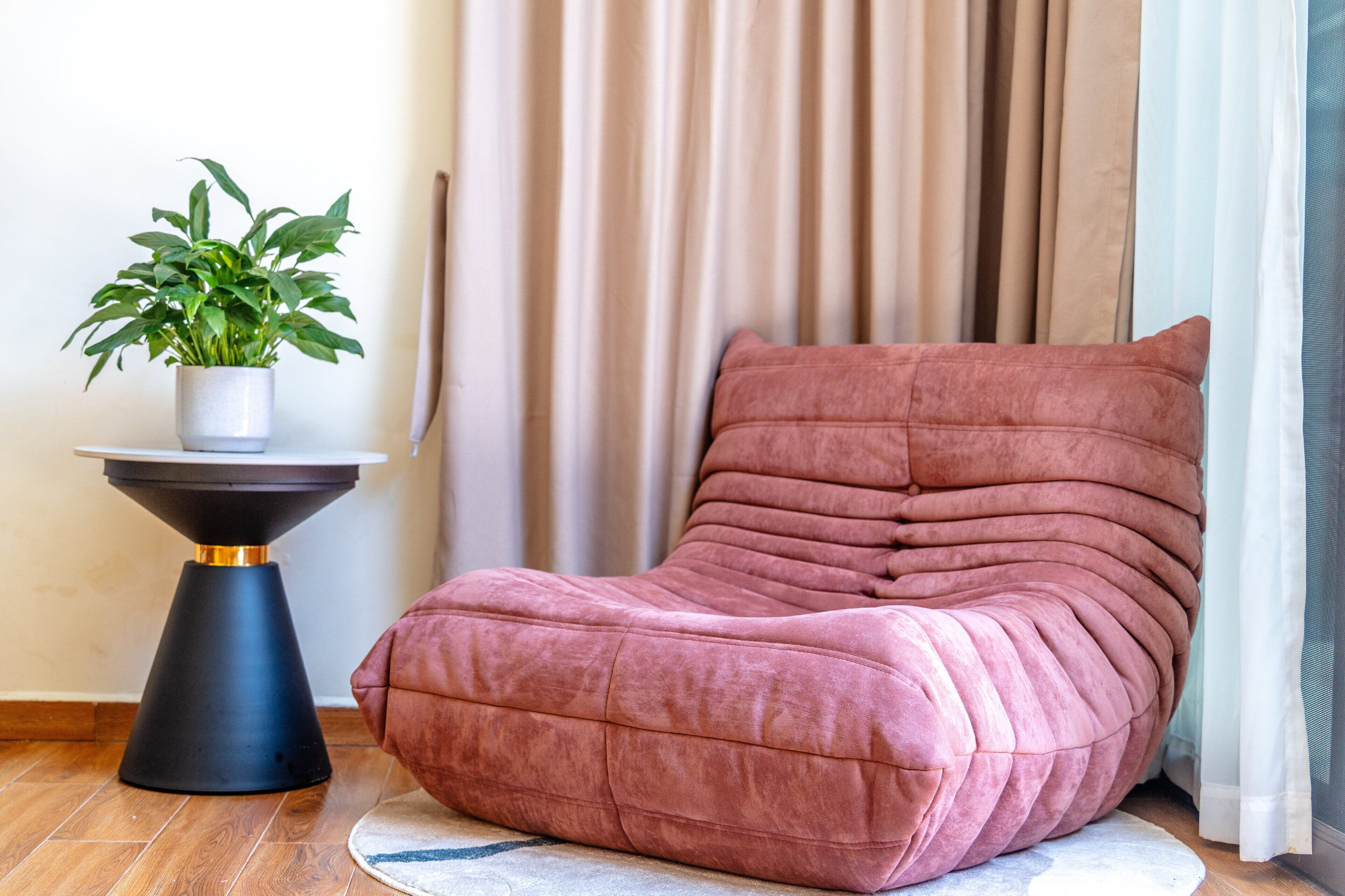 A pink cushioned chair with a round table beside it, a potted plant, and a curtain in the background.