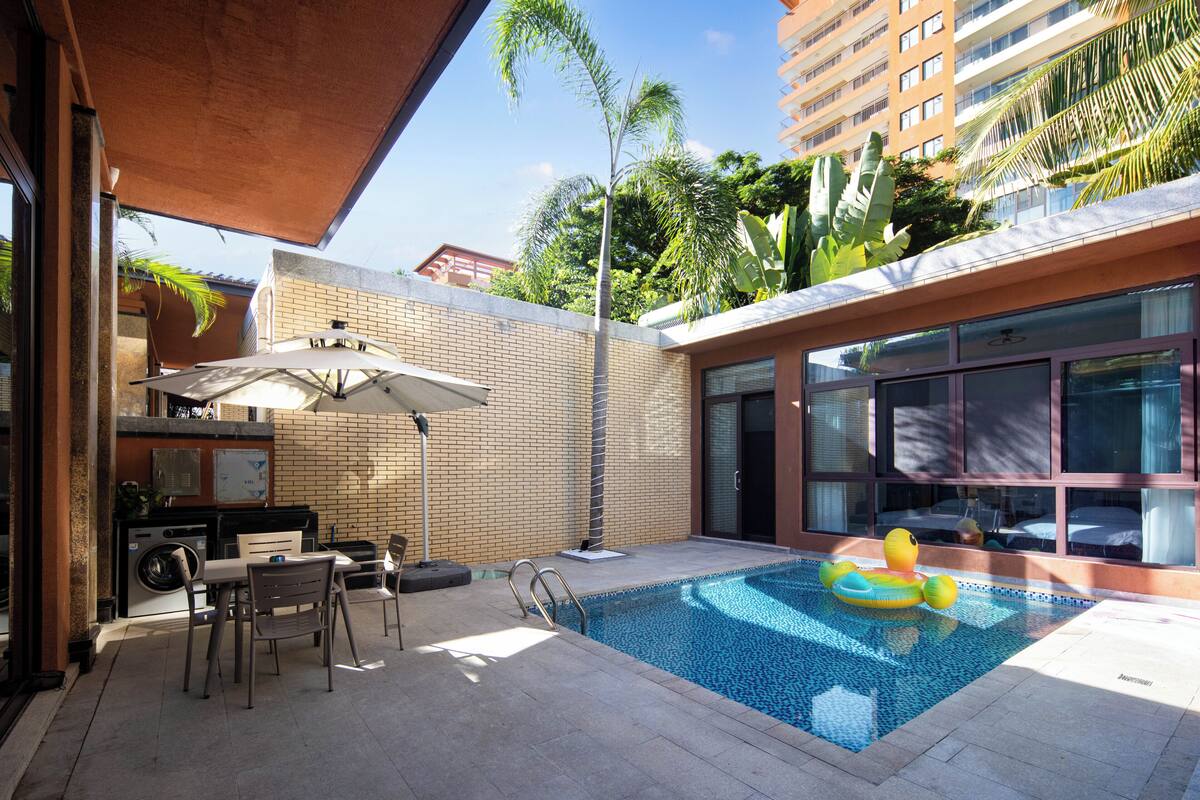 A swimming pool with a lounge area, a covered seating area, and a view of a tall building in the background.