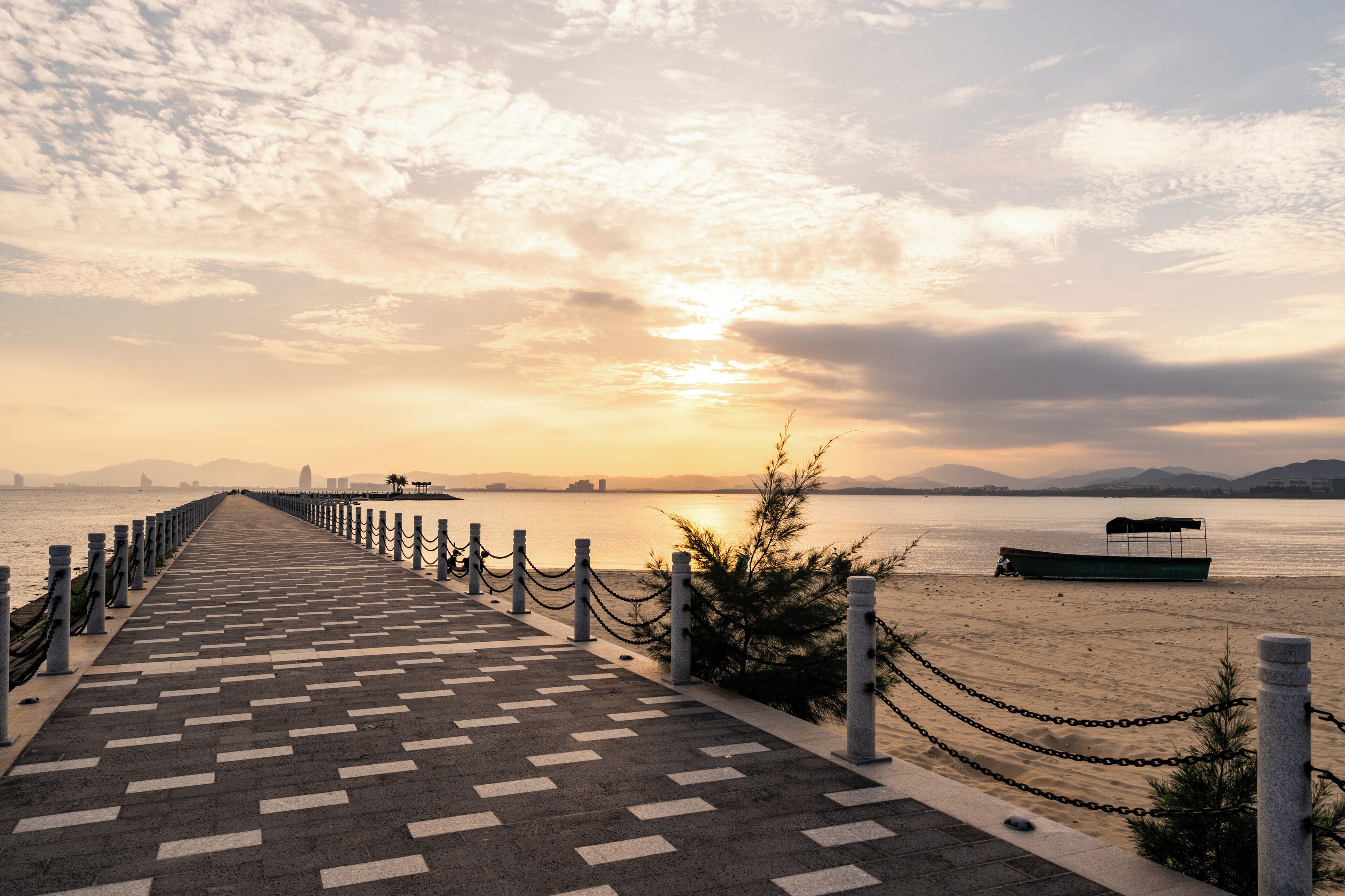 Ein gepflasterter Weg führt zu einem Strand, auf dem ein Boot liegt, mit einer entfernten Stadtlandschaft im Hintergrund.