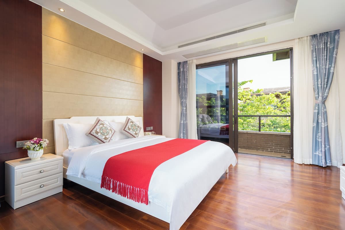 A modern bedroom with a large bed, a nightstand, and a view of greenery through a sliding glass door.