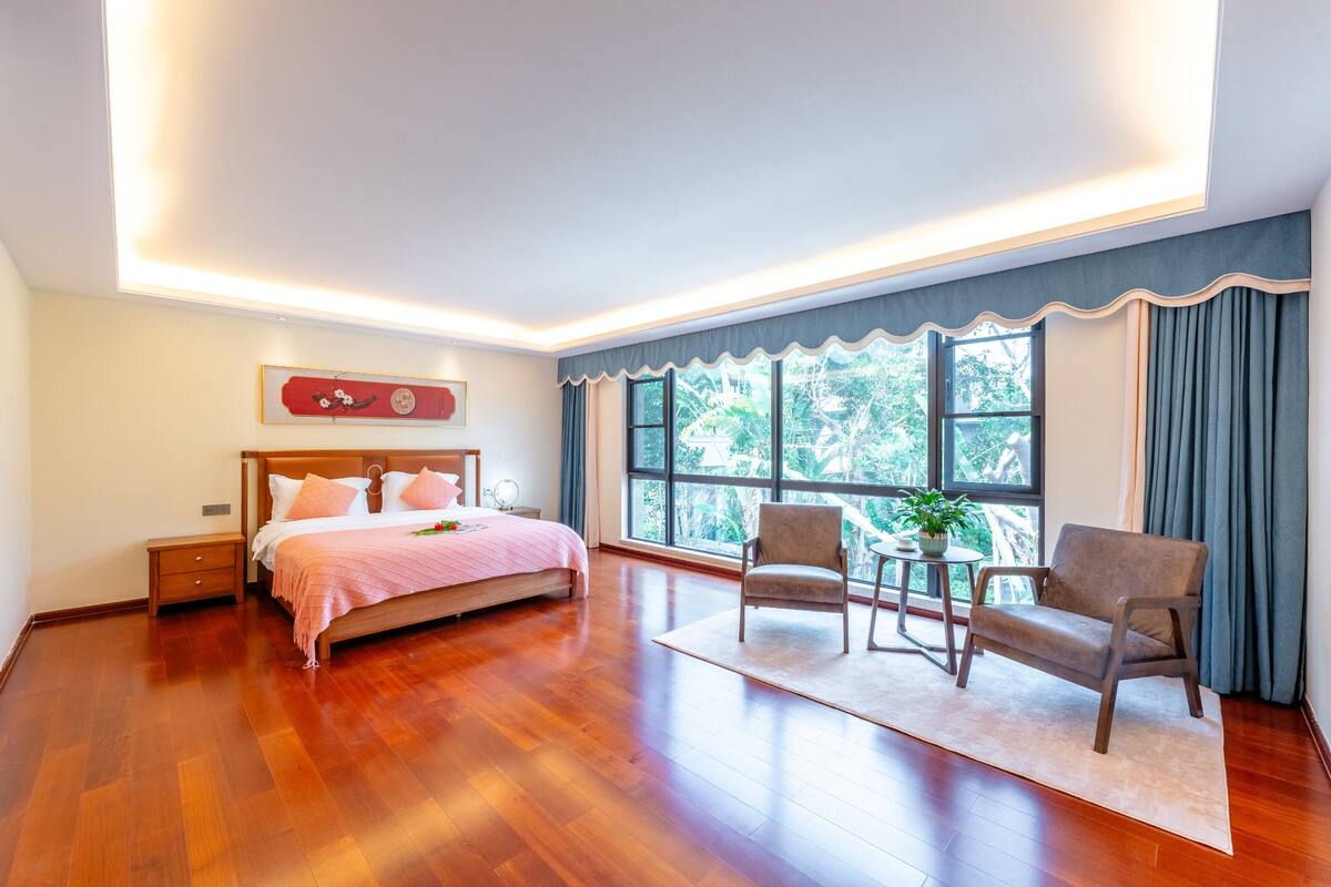 A bedroom with a large bed, a nightstand, a chair, a small table with a plant, and a window with curtains.