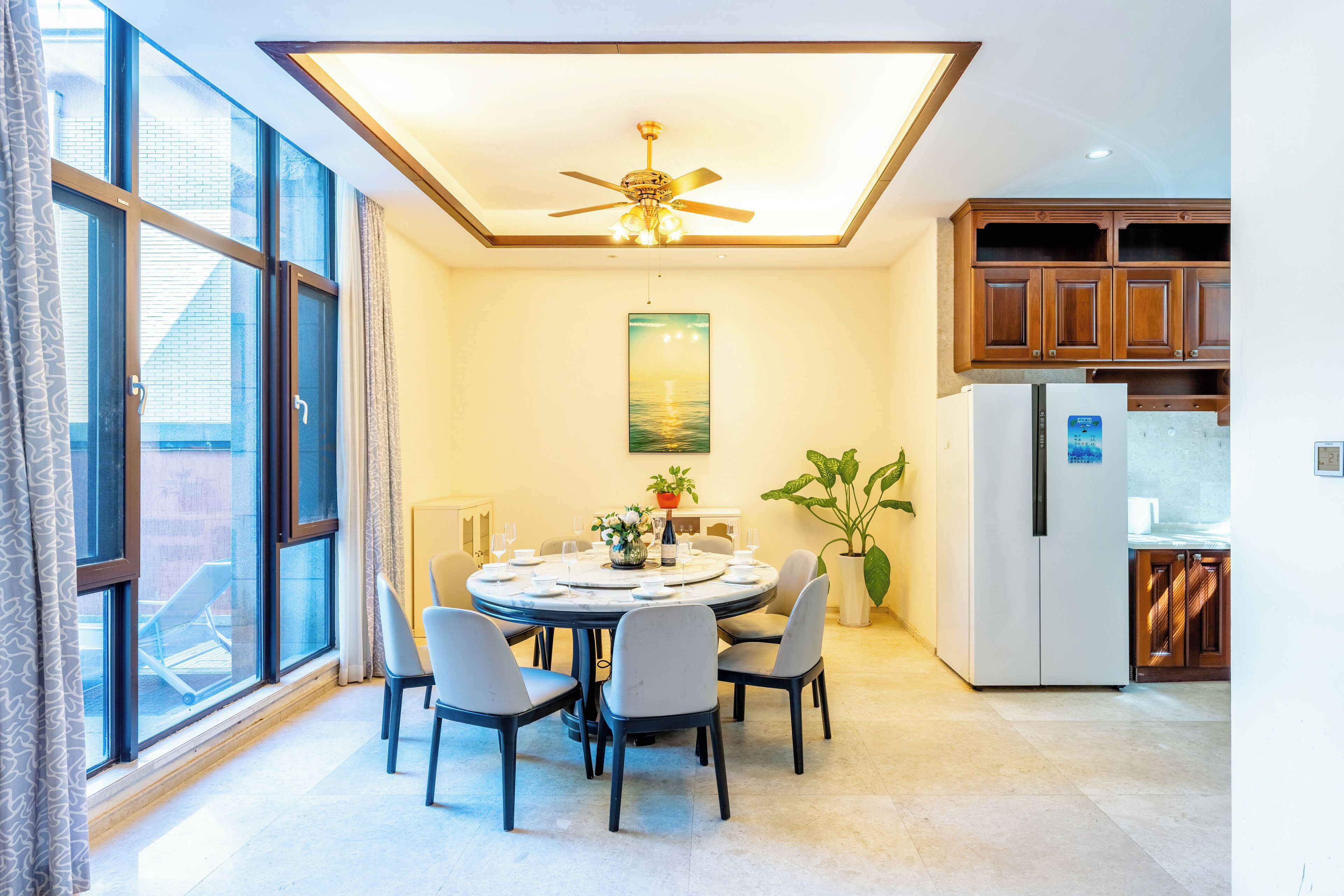 A dining area with a round table, chairs, a ceiling fan, and a view through large windows.