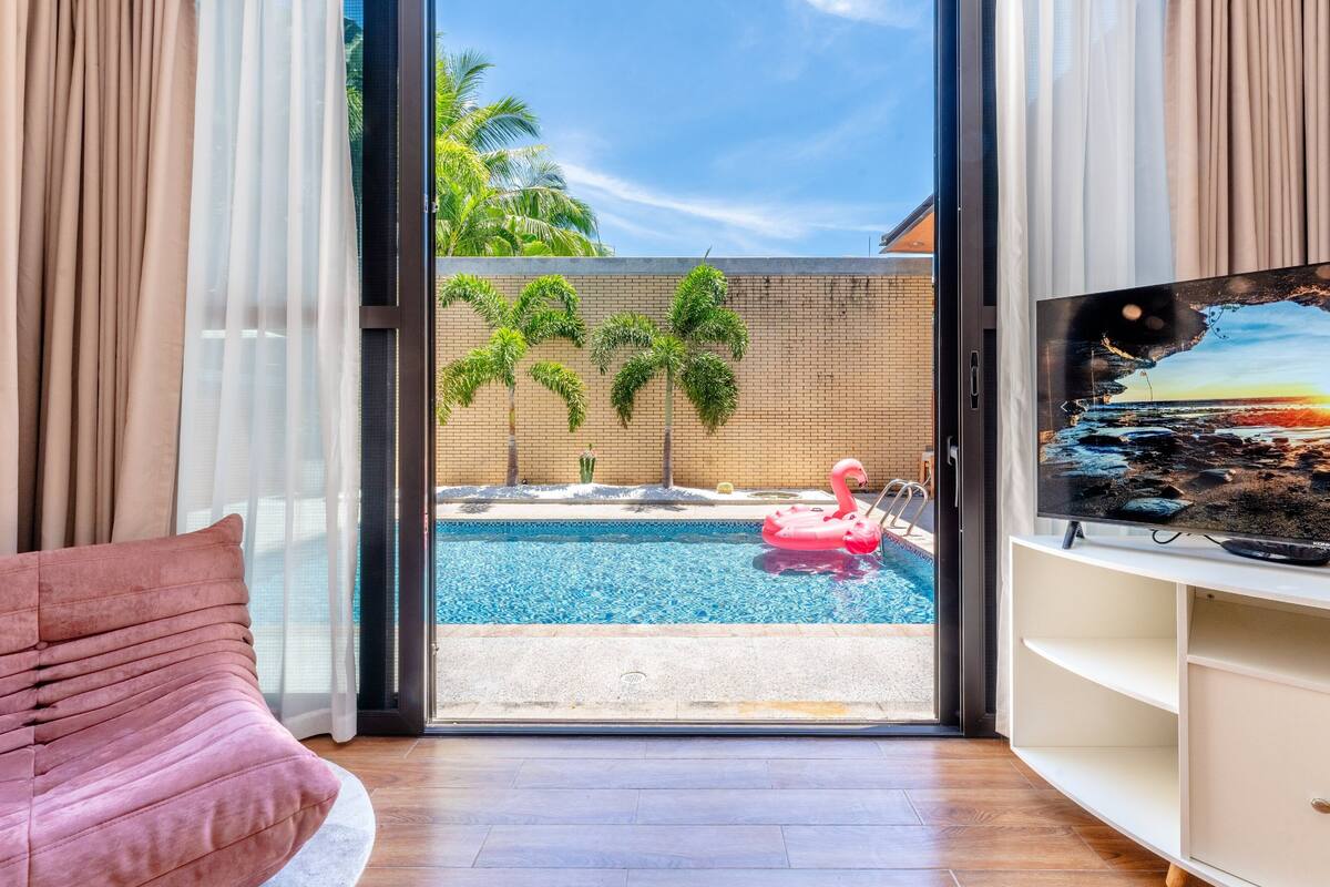A modern living room with a large window overlooking a swimming pool, a pink flamingo float, and a TV on a white shelf.