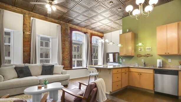 Una sala de estar moderna con sofá, sillón y mesa de centro. Se ve una cocina con armarios de madera y una zona de comedor con sillas.