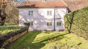 A two-story house with a white exterior, a brick chimney, and a tiled roof. There is a small garden with a lawn, a hedge, and a table with chairs. - Mulberry End, Foxhall (Foxhall)
