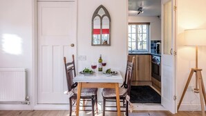 A dining area with a table set for two, a window with a view, and a kitchen visible through an open door. - Mulberry End, Foxhall (Foxhall)