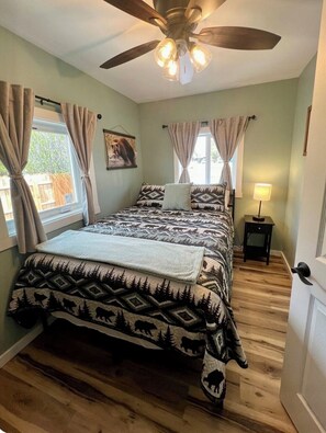 Une chambre à coucher avec un lit, un ventilateur de plafond, une fenêtre avec des rideaux, une table de chevet et une porte.