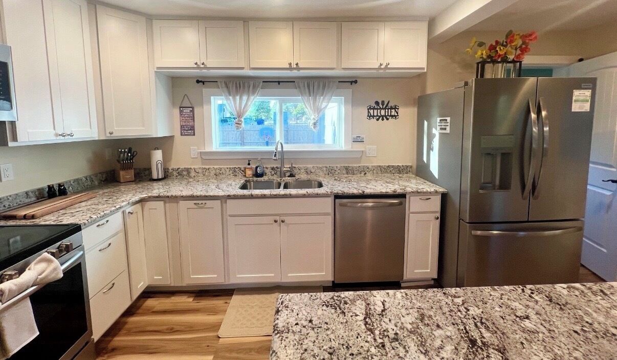Una cocina con armarios blancos, un refrigerador de acero inoxidable, una encimera de granito y una ventana con cortinas.