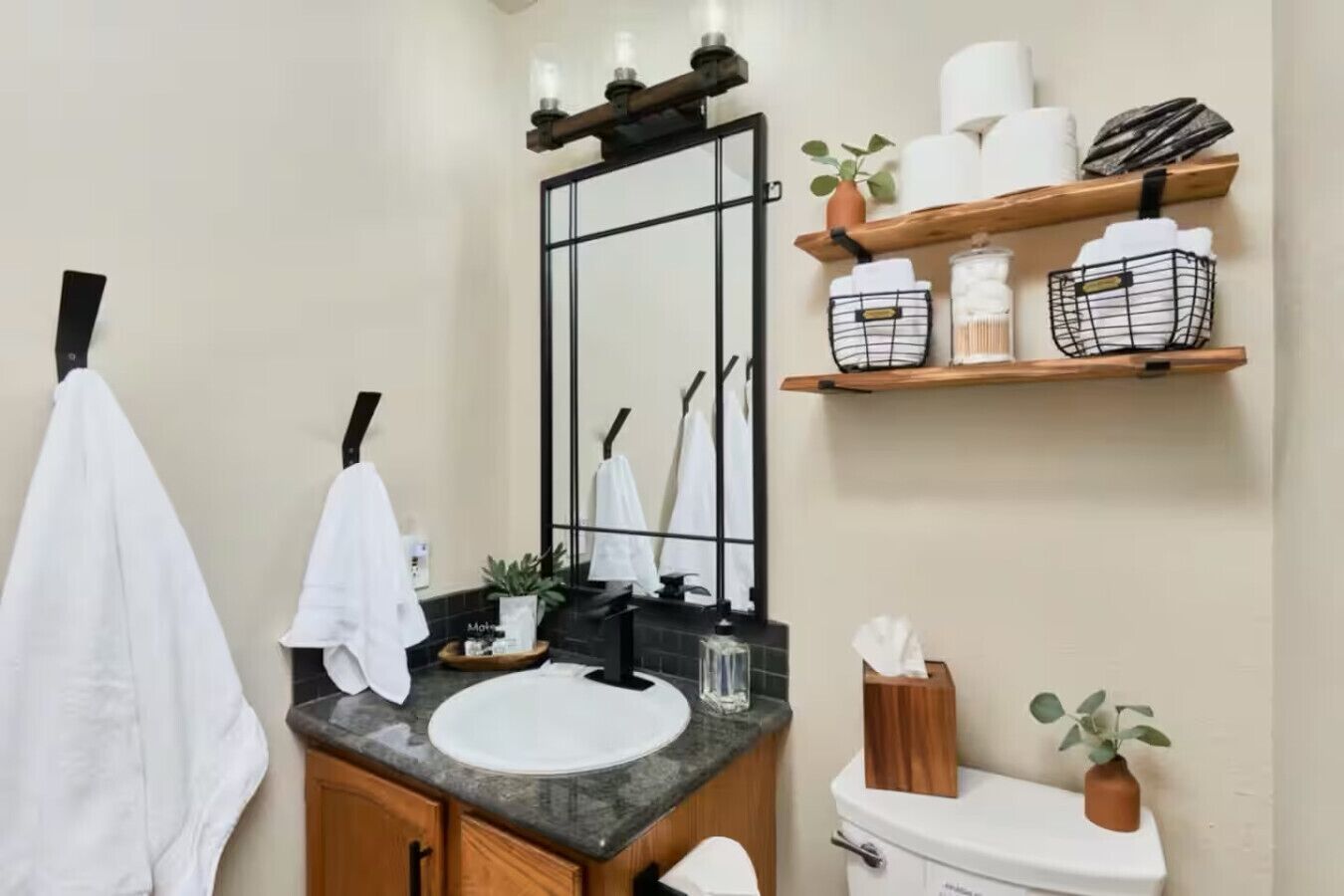A bathroom with a white sink, black countertop, wooden cabinets, a mirror with black frame, and a toilet. There are towels hanging on a rack, a plant on the countertop, and shelves with toiletries.