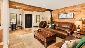 A cabin interior with a wooden wall, a brown leather sofa, a wooden coffee table, and a fireplace. - Family Cabin • Hot Tub • Disc Golf • Forest Access (Divide)