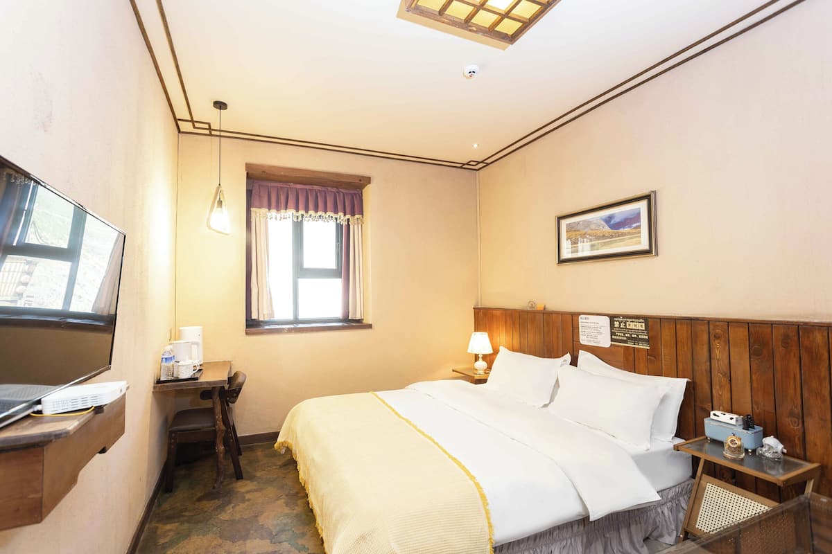 Een hotelkamer met een bed, een bureau met een computer, een lamp, een raam met gordijnen, en een schilderij aan de muur.