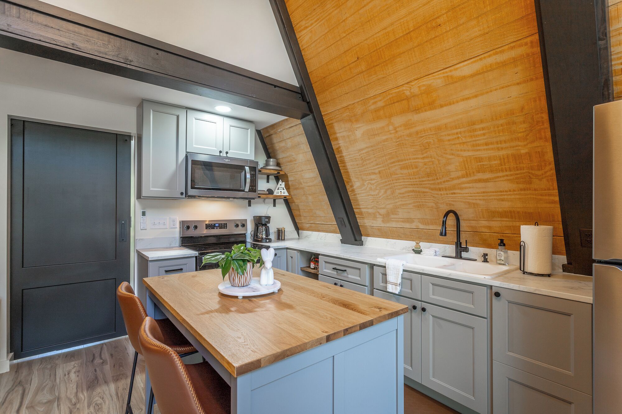 A modern kitchen with a wooden ceiling, white cabinets, a built-in microwave, and a wooden island with a chair.