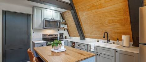 A modern kitchen with a wooden ceiling, white cabinets, a built-in microwave, and a wooden island with a chair.