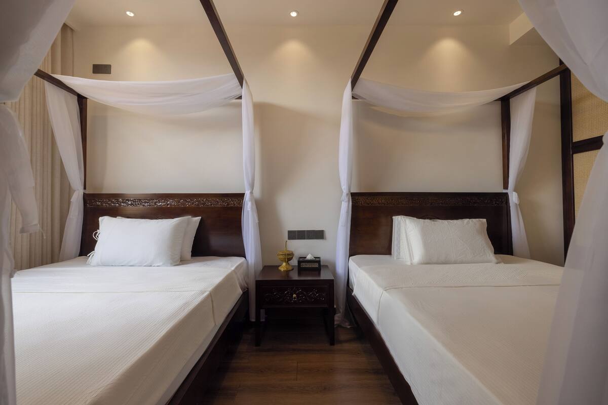 A hotel room with two beds, each with white linens and a wooden headboard. There is a nightstand between the beds with a lamp and a small table.