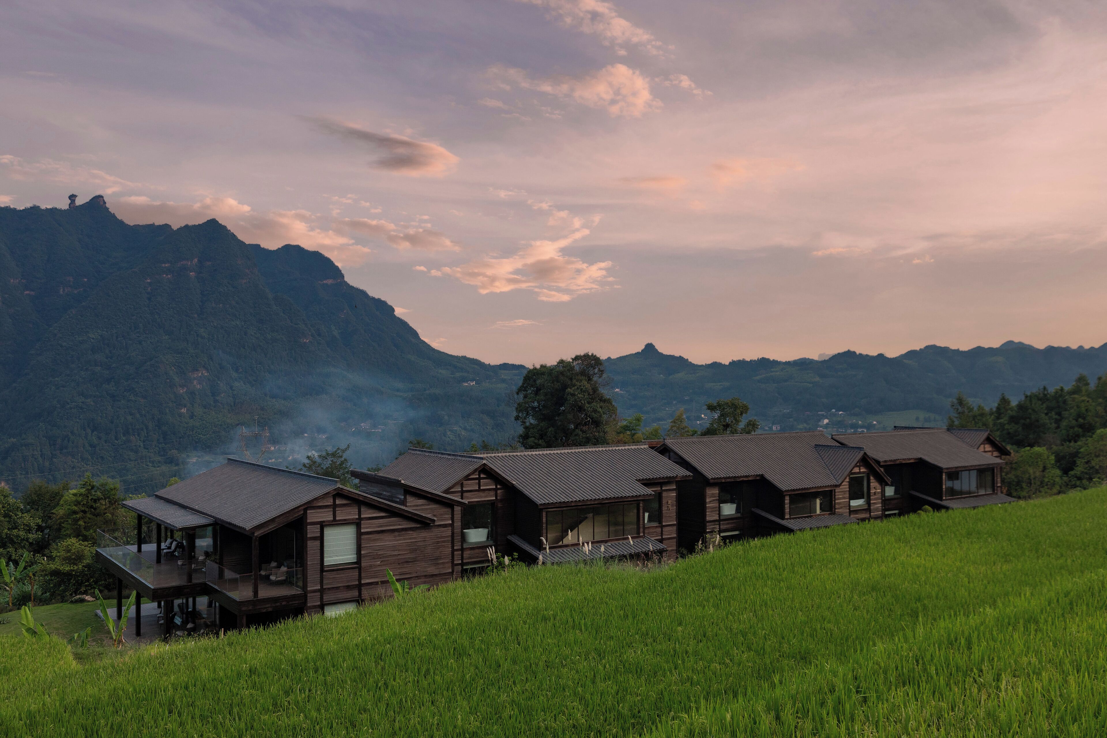 A row of wooden cabins with large windows and balconies, set against a backdrop of mountains and a cloudy sky.