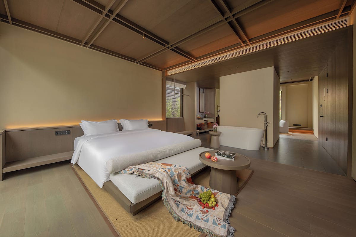 Une chambre d’hôtel moderne avec un grand lit, une table d’appoint ronde sur laquelle se trouve un panier de fruits, une baignoire et un petit coin salon.