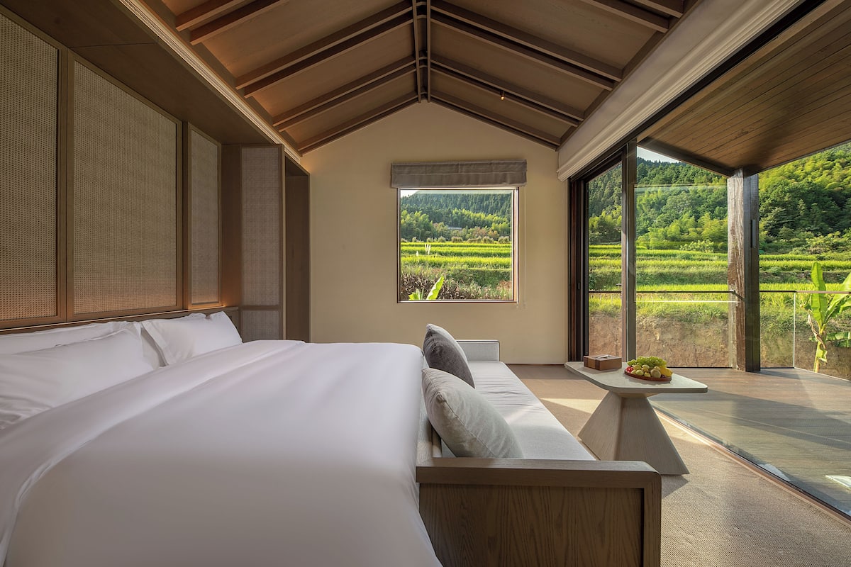 Une chambre avec un grand lit, une petite table sur laquelle sont disposés des fruits, et une vue sur un paysage verdoyant par la fenêtre.