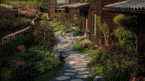 A wooden house with a stone pathway leading to it, surrounded by lush greenery and a mountainous backdrop. - Country Retreats Zhangjiajie Manson Resort (Zhangjiajie)