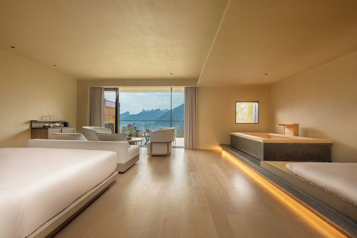 Une chambre d’hôtel moderne, dotée d’un grand lit, d’un canapé, d’une petite table et offrant une vue sur les montagnes grâce à une grande fenêtre.