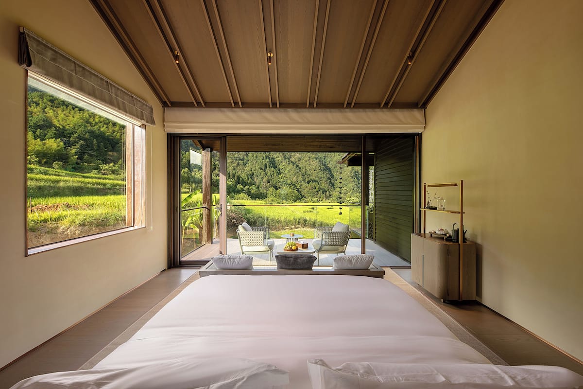 Une chambre avec un grand lit, une vue sur un paysage verdoyant, une petite table avec des fruits et un coin salon avec des chaises.