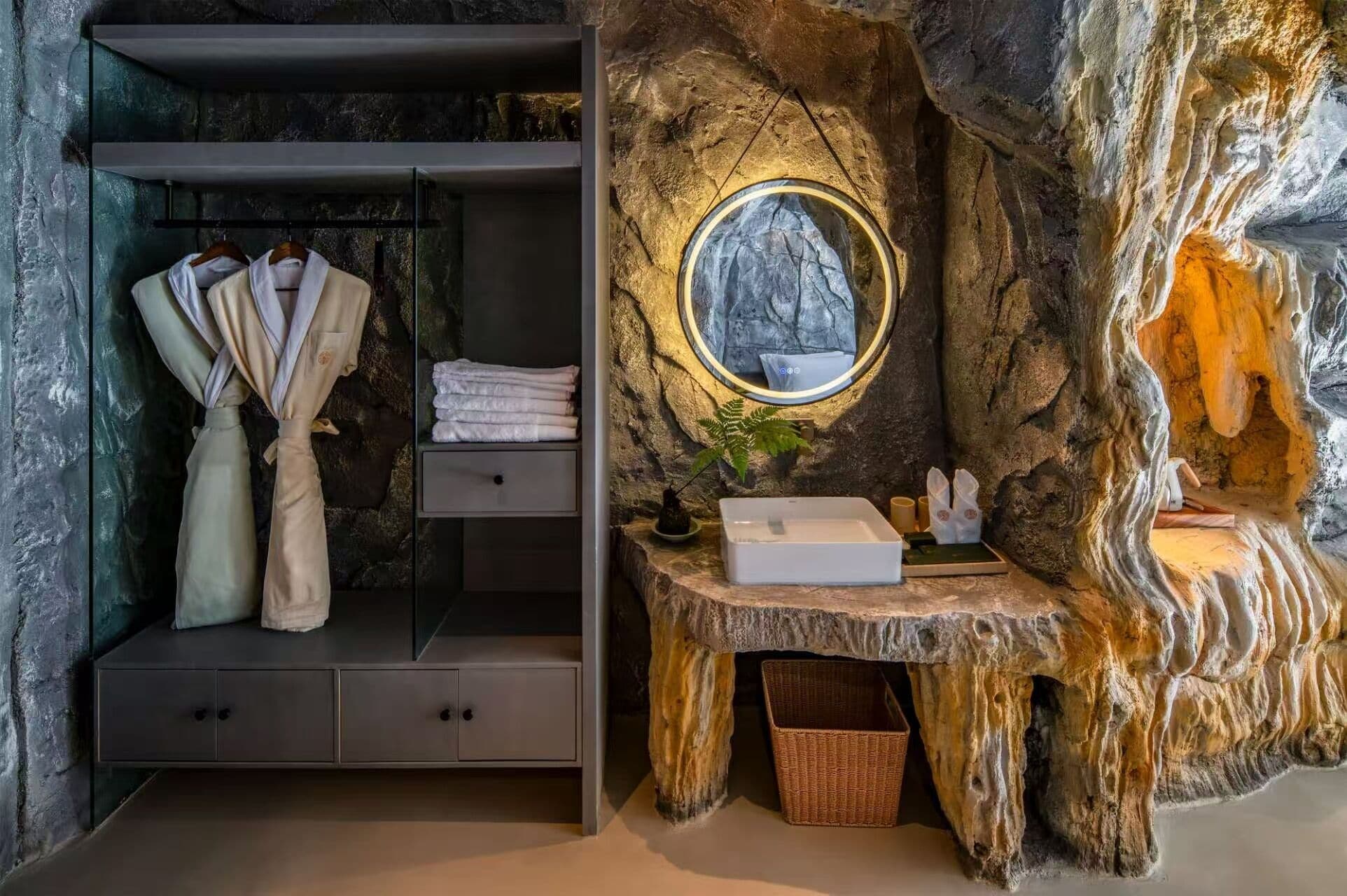 A bathroom with a rustic stone wall, a wooden vanity with a sink, and a robe rack with towels.