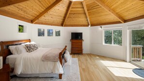 Une chambre avec un plafond en bois, un lit, une commode, un téléviseur et de grandes fenêtres.