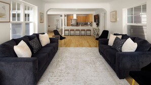 Un salon avec deux canapés sombres, un tapis à motifs clairs et une cuisine à l’arrière-plan.