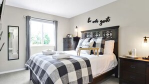 A bedroom with a checkered bedspread, a bedside table with a lamp, a dresser, and a wall sign saying "be our guest".