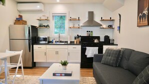 Eine kompakte Küche mit Kühlschrank, Mikrowelle, Herd und Spüle. Es gibt einen Essbereich mit Tisch und Stühlen sowie ein graues Sofa im Wohnzimmer.