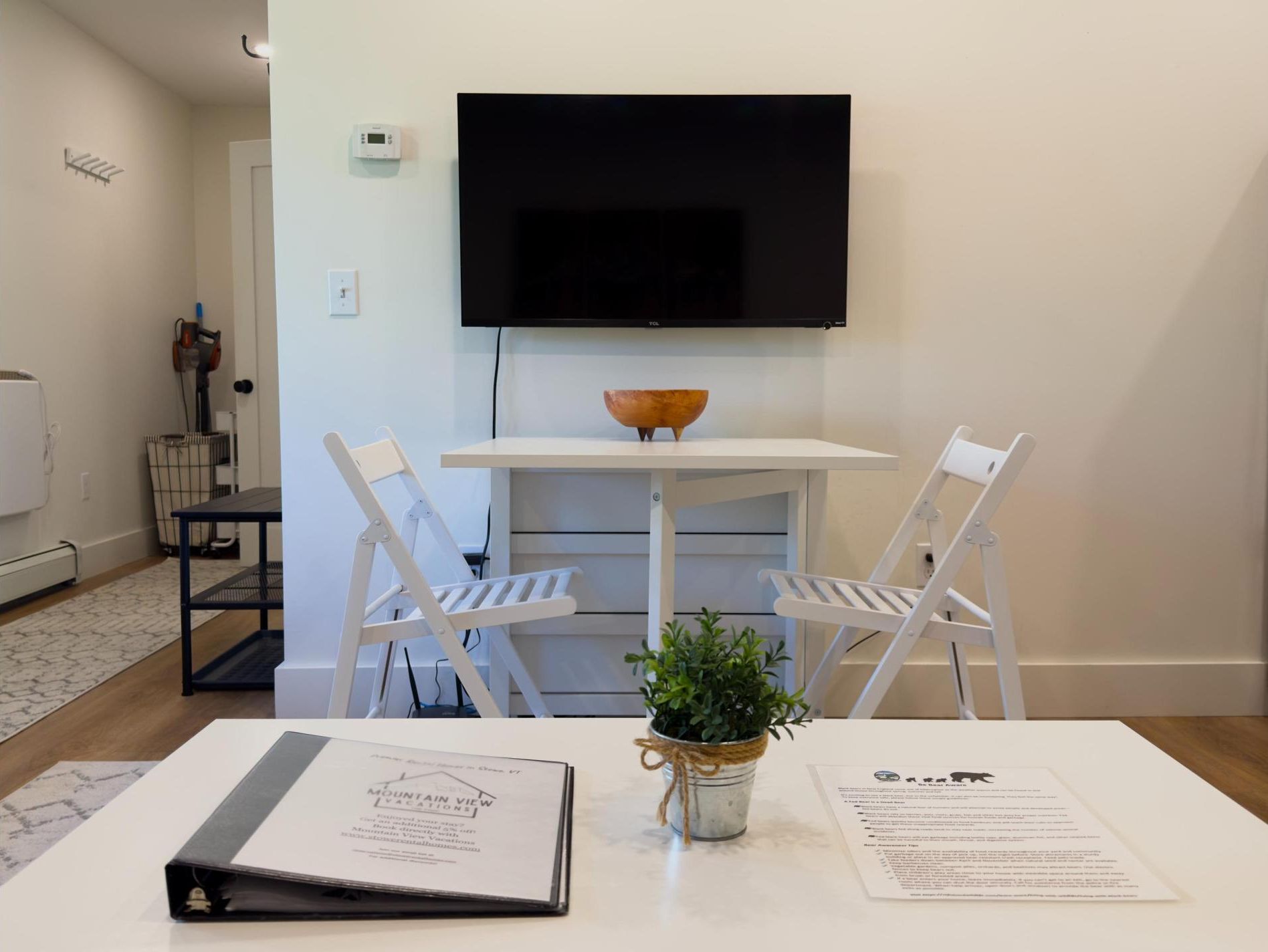 Een moderne woonkamer met een flatscreen televisie aan de muur, een witte tafel met een schaal en een bloempot, twee witte stoelen en een witte bank.