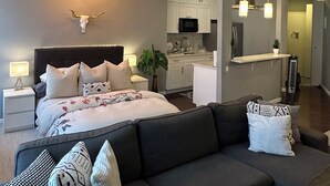 Una sala de estar moderna con un sofá gris oscuro, una cama grande con ropa de cama blanca, una araña y una cocina con gabinetes blancos y un microondas.