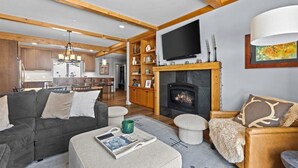 A cozy living room with a fireplace, a television, and a dining area in the background.