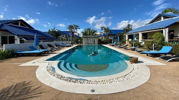 A swimming pool with lounge chairs and umbrellas, surrounded by buildings with blue roofs and palm trees.