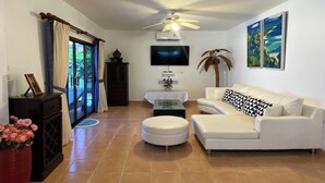 A living room with a white sofa, a ceiling fan, a flat-screen TV, a small table with a vase, and a large potted plant.