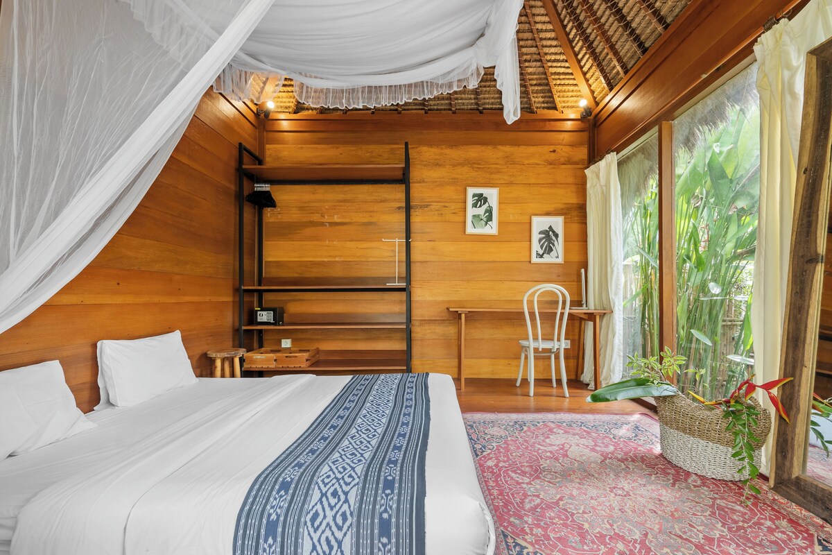 A bedroom with a canopy bed, wooden walls, a desk, and a large window with a view of greenery.