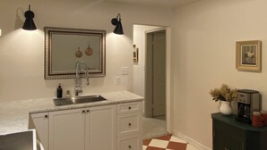 Una cucina con pavimento a scacchi, mobili bianchi, un lavello e uno specchio sopra. Si nota una luce a soffitto e una macchina da caffè su un mobile.