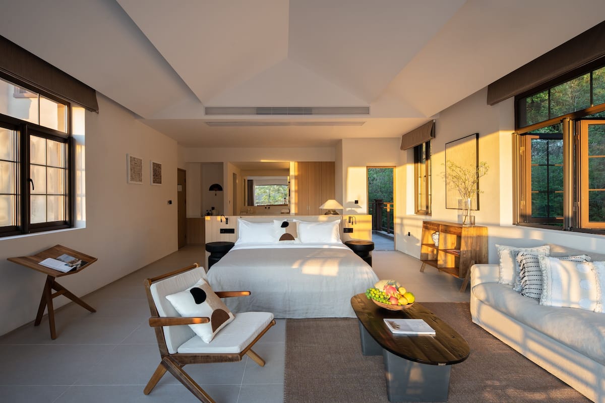 A modern hotel room with a large bed, a sofa, a wooden coffee table, and a small table with a bowl of fruit.