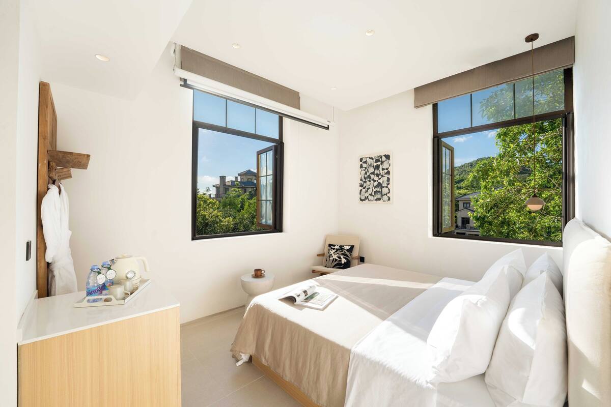 A modern bedroom with a large bed, a bedside table, a small bedside table, a window, and a view of greenery outside.