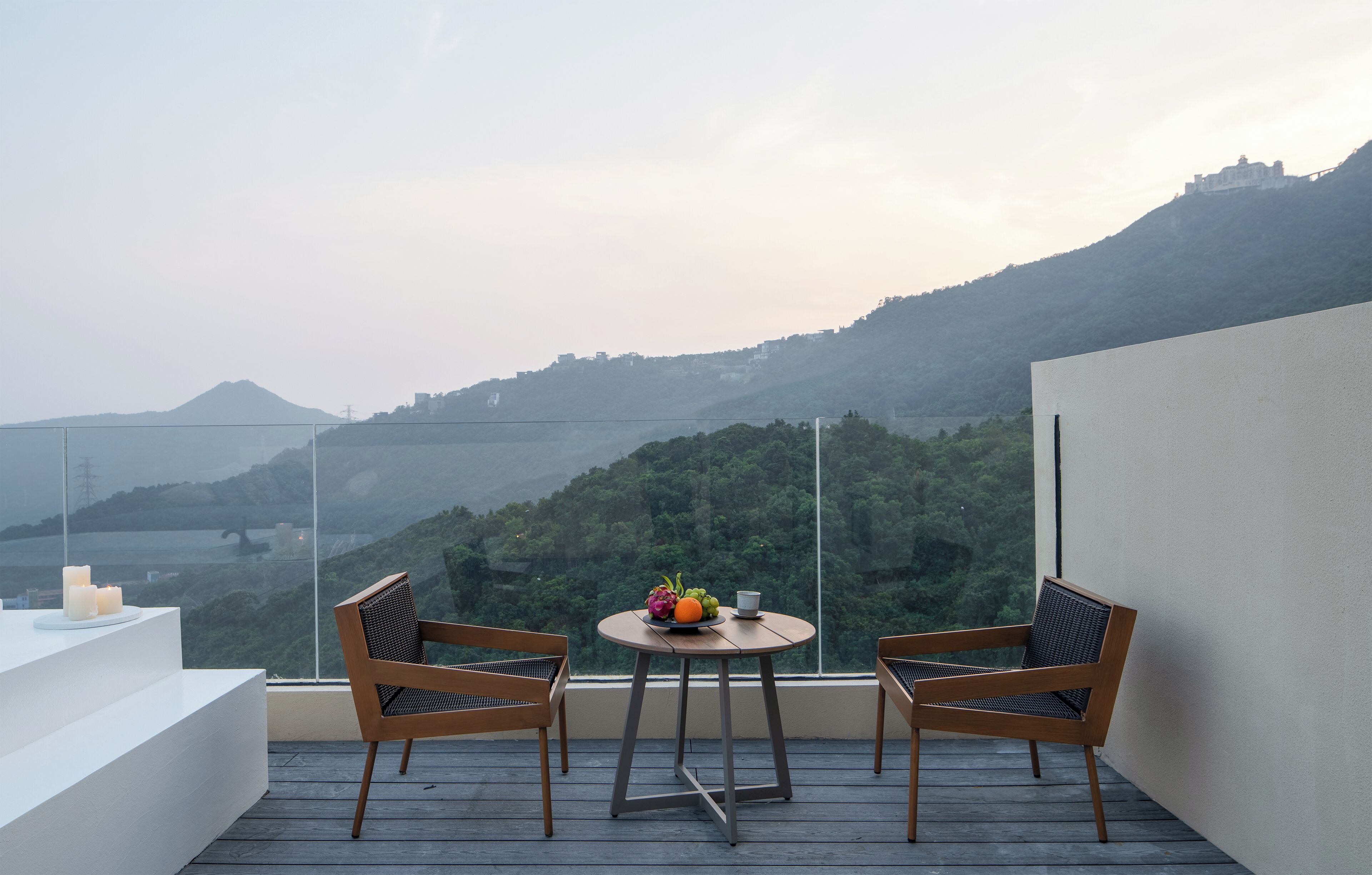 A balcony with two wooden chairs, a small round table with a fruit bowl, and candles. The balcony overlooks a mountainous landscape with a view of a castle-like structure on a hill.