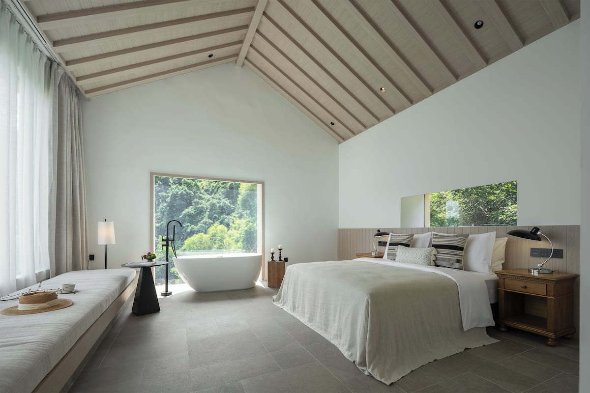 A modern bedroom with a large window, a bathtub, a bed, and a bedside table.