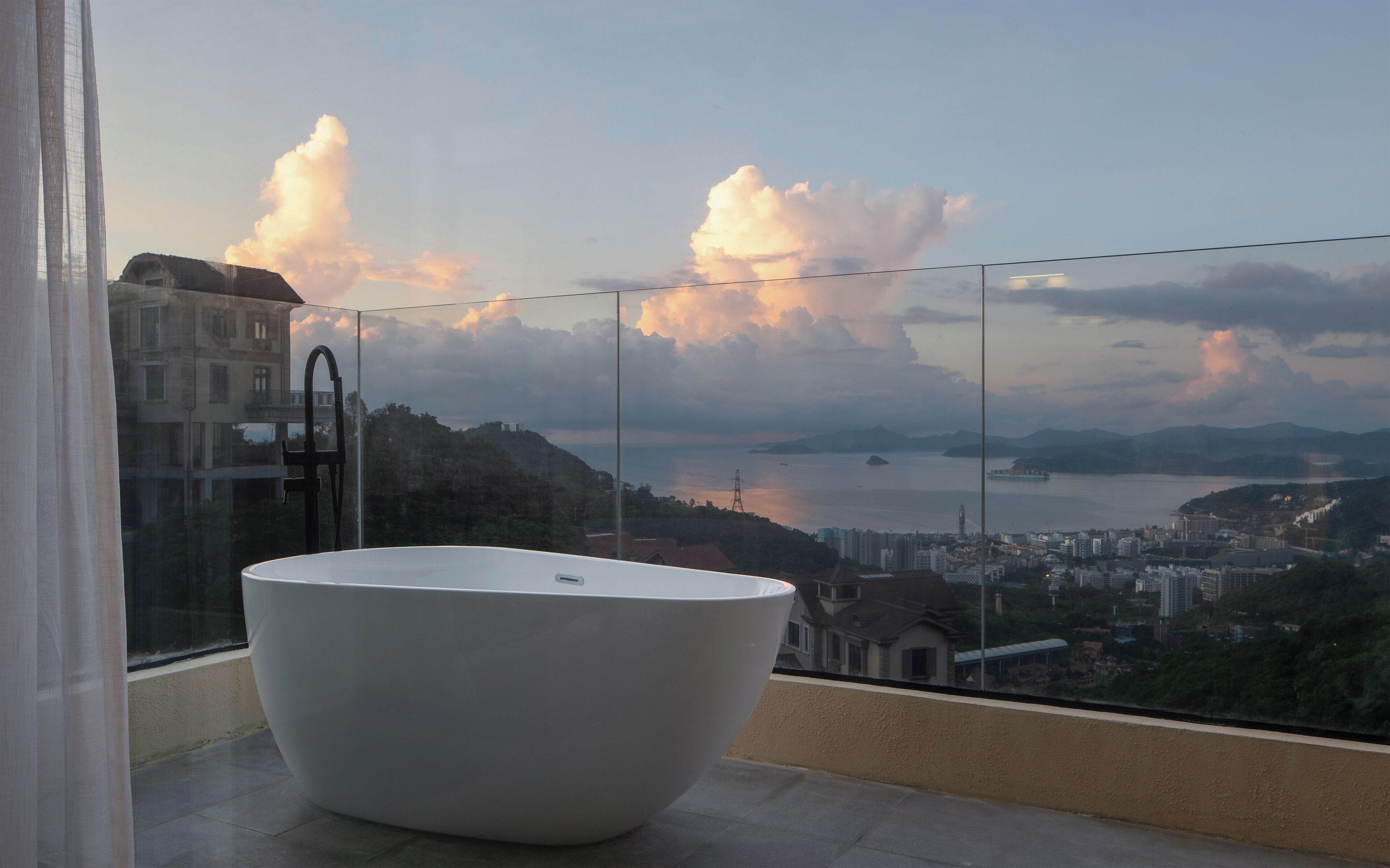 A large white bathtub in a balcony area with a cityscape view, including mountains and a body of water.