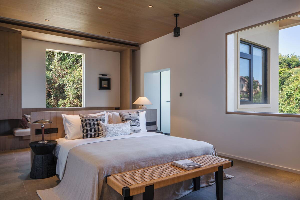A modern bedroom with a large bed, a bedside table, a lamp, and a window with a view of greenery.