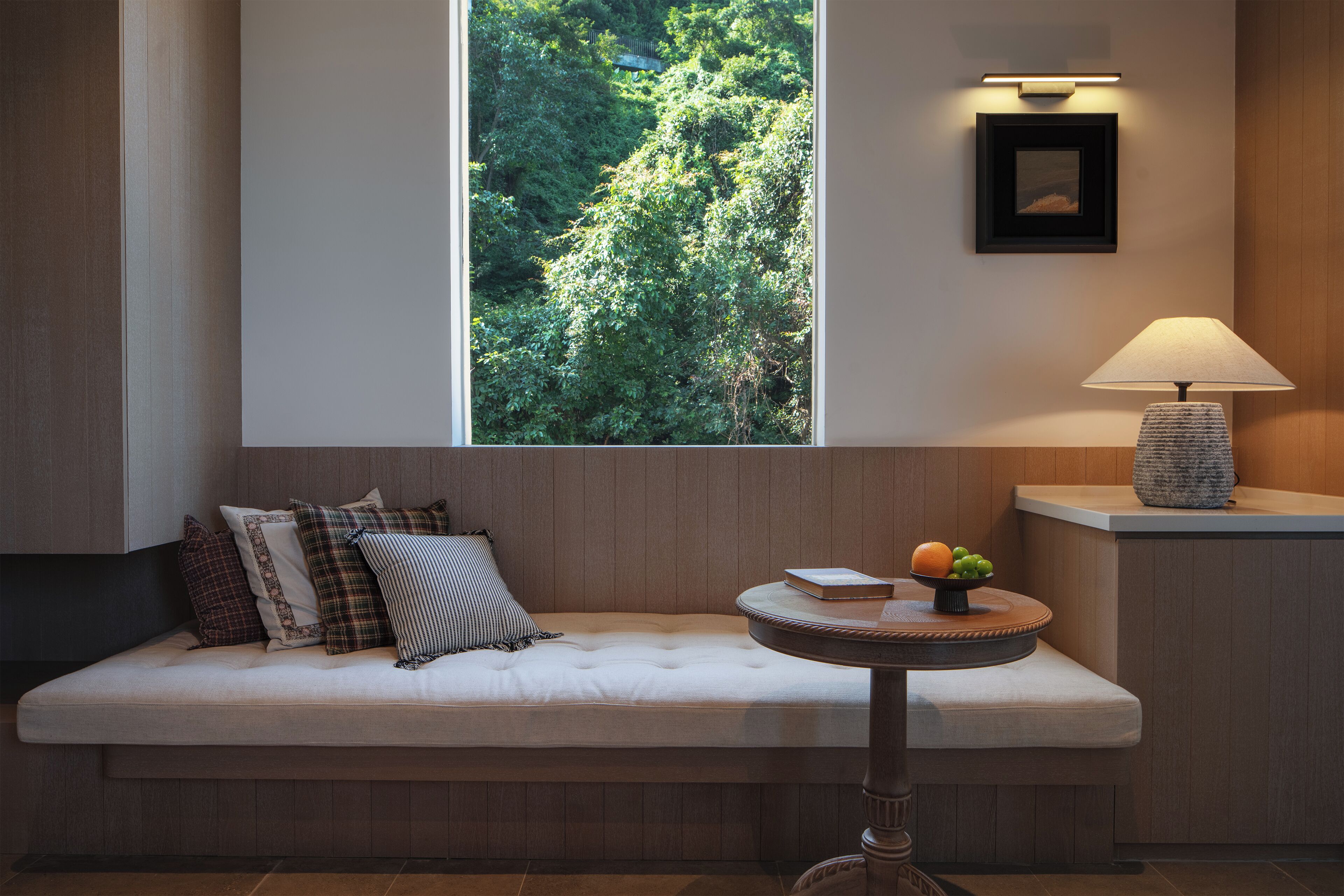 Et moderne værelse med en træbænk, et rundt bord med en lampe og et vindue med udsigt til grønne omgivelser.